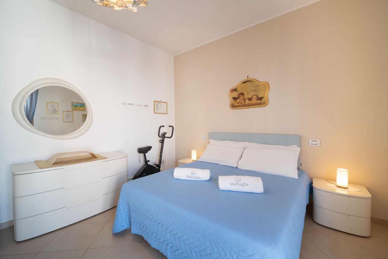A bedroom with a bed, a bedside table, a nightstand, a mirror, and a wall-mounted picture.