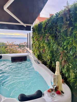 Un jacuzzi con una botella de champán y fresas, rodeado de exuberante follaje y vistas al mar.