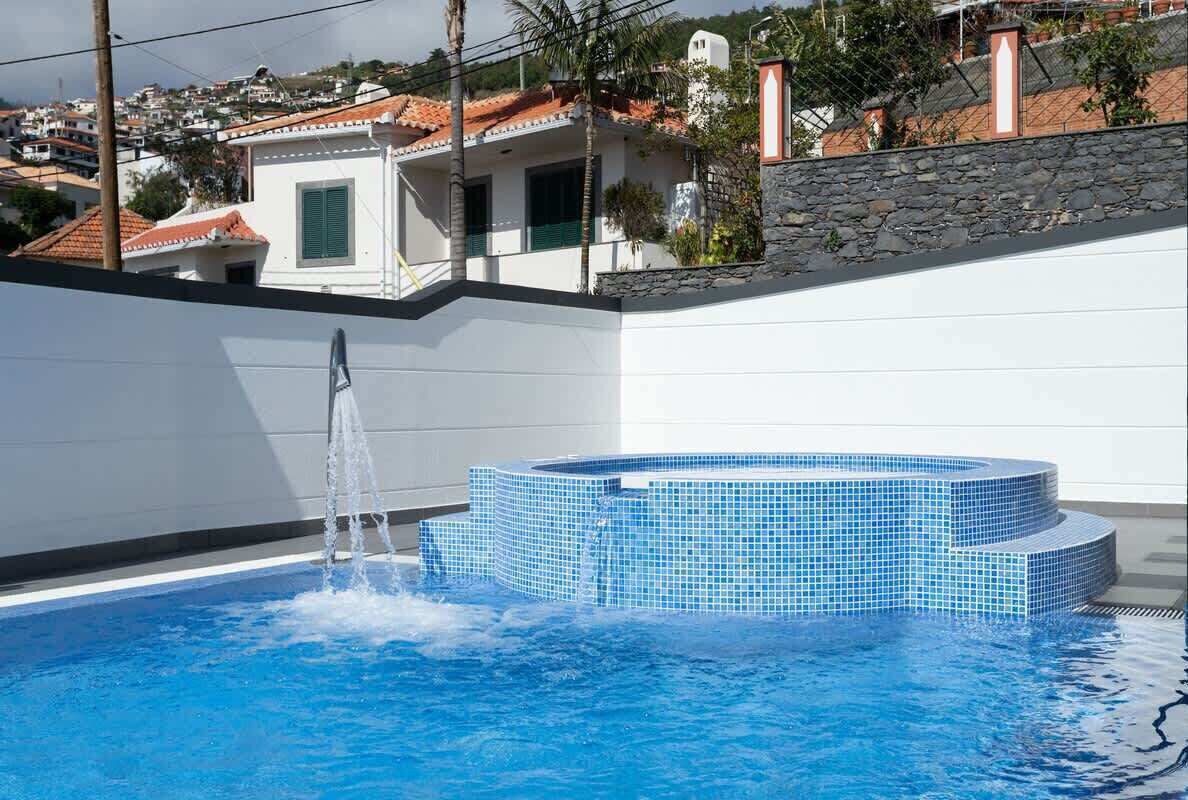 Una piscina con fuente, un muro de piedra y un edificio blanco con tejado de tejas al fondo.
