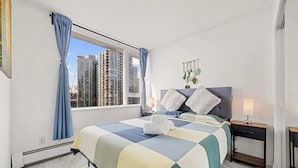 Hotelkamer met een groot bed, nachtkastje, hangplant en uitzicht op hoge gebouwen.