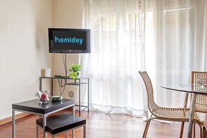 A room with a television displaying 'homiday,' a small table with a plant, a wicker chair, and a glass table.