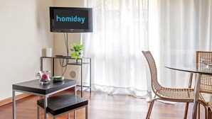 Una stanza con una TV che mostra "homiday", un piccolo tavolo con una pianta, sedie in vimini e una panca.