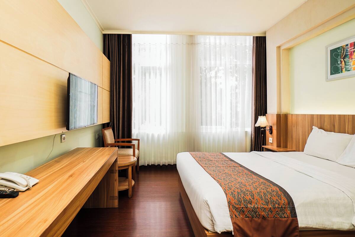 Une chambre d’hôtel avec un bureau en bois, un lit recouvert d’une couvre-lit à motifs, un téléviseur fixé au mur et une fenêtre ornée de rideaux légers.