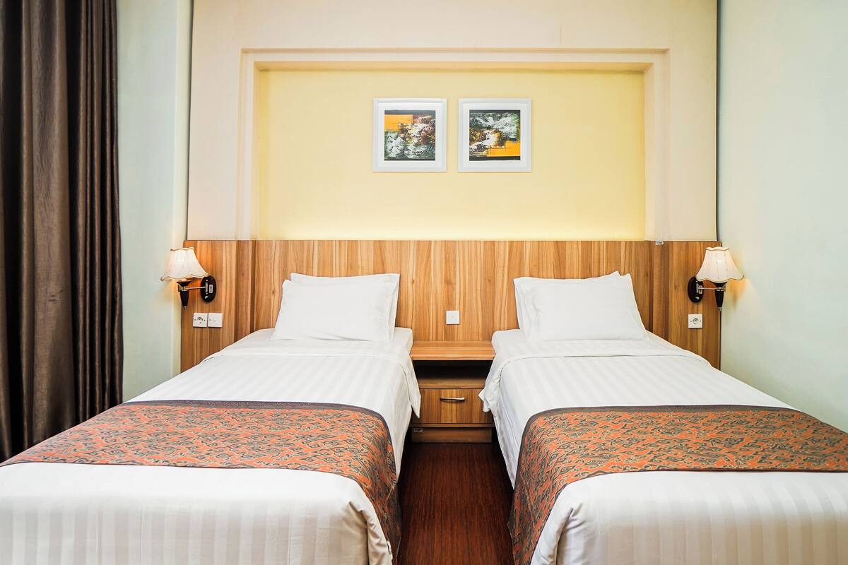 Une chambre d’hôtel avec deux lits simples, une tête de lit en bois, deux lampes fixées au mur et deux tableaux encadrés accrochés au mur.