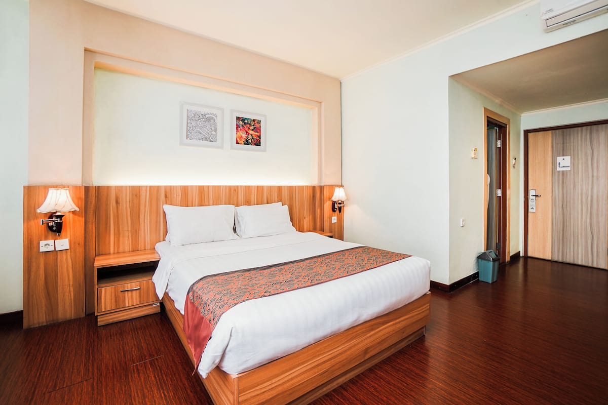 Une chambre d’hôtel avec un lit en bois, deux tables de chevet avec des lampes, un petit tiroir et une étagère fixée au mur.