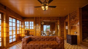 A log cabin interior with a tufted leather sofa, a dining table with chairs, and a ceiling fan. - Villa 107 Amarja's Citadel Nest (Mawal)