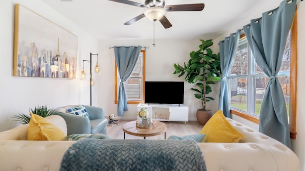 Ein Wohnzimmer mit einer Couch, einem Couchtisch, einem Fernseher, einem Deckenventilator und einem großen Fenster mit Vorhängen.