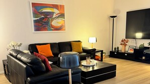 Een moderne woonkamer met een zwarte hoekbank, een salontafel, een vloerlamp en een flatscreen televisie die aan de muur is bevestigd.