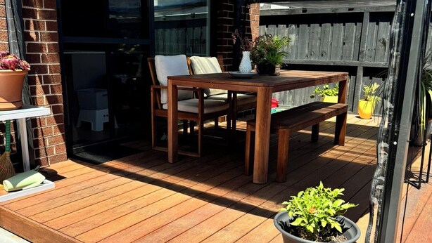 Een houten terras met een tafel, stoelen, potplanten en een overdekt gedeelte met een raam.