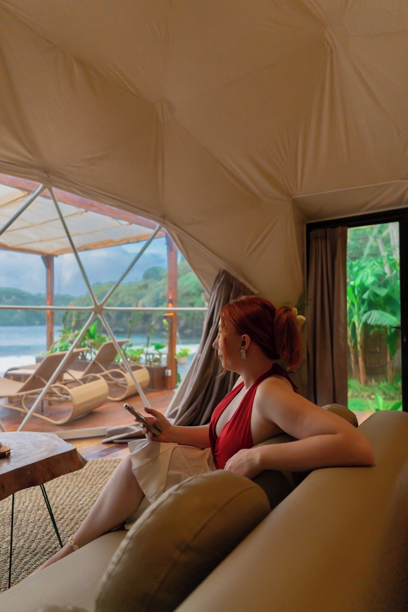 A person sitting on a sofa inside a tent-like structure with a view of a body of water and greenery outside.