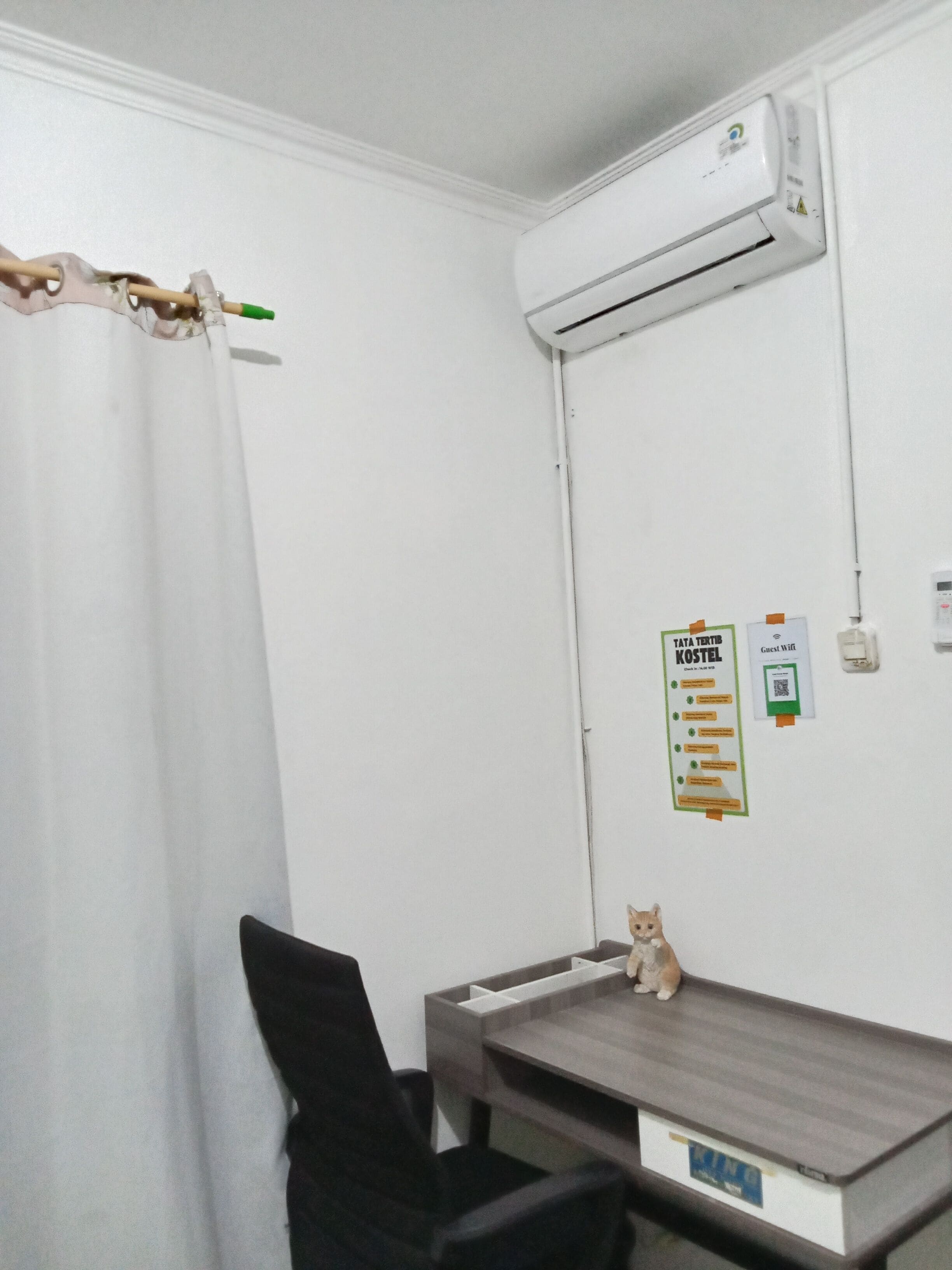 A room with a white air conditioner, a curtain, a desk with a cat, and a wall with a sign and a QR code.