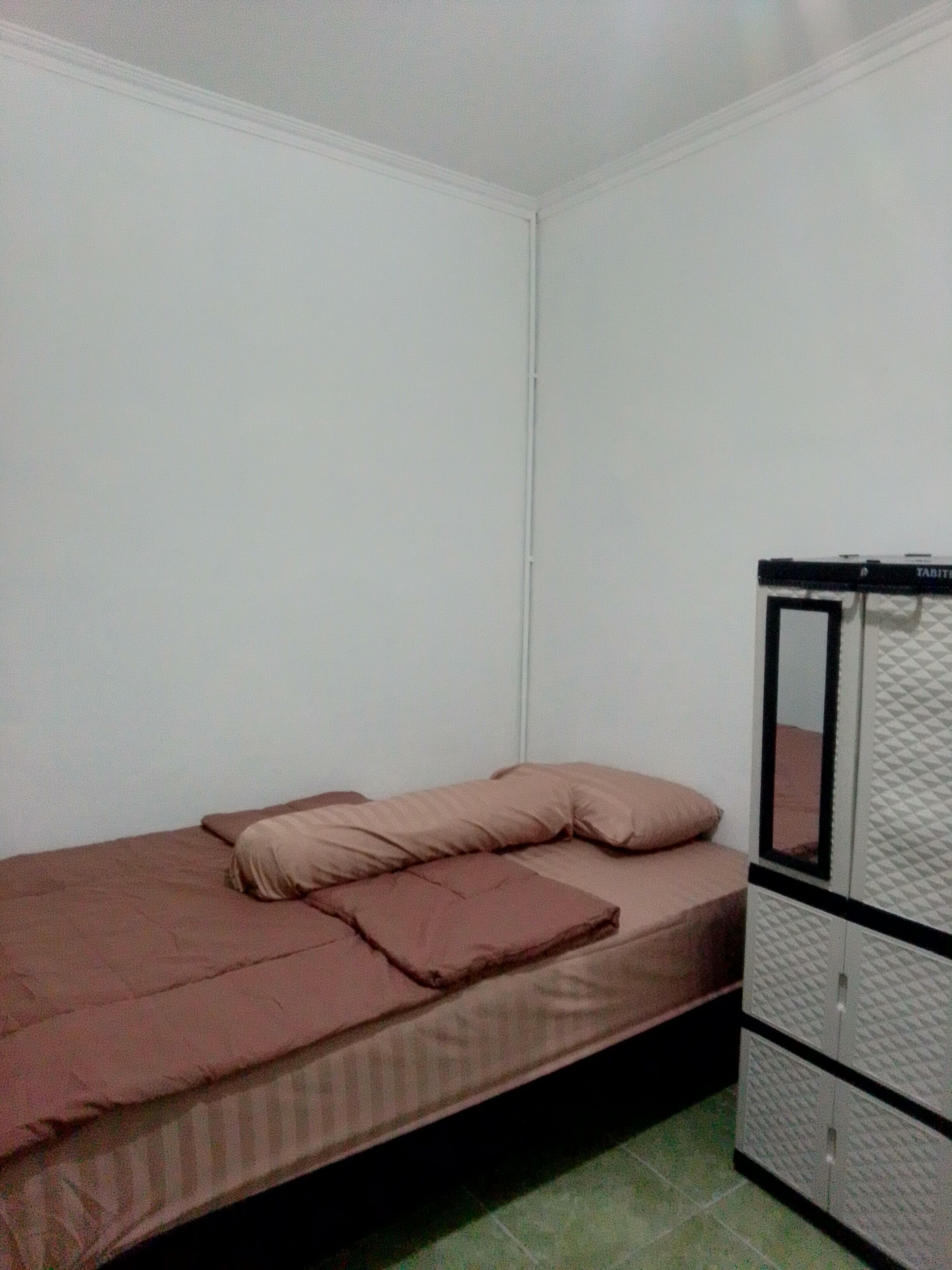 Una stanza piccola con un letto singolo, un muro bianco semplice e un pavimento in piastrelle verdi.