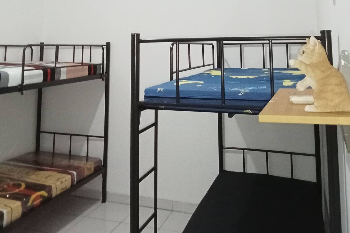 Een studentenkamer met stapelbedden, een klein tafeltje en een luchtverkoeler aan de muur.