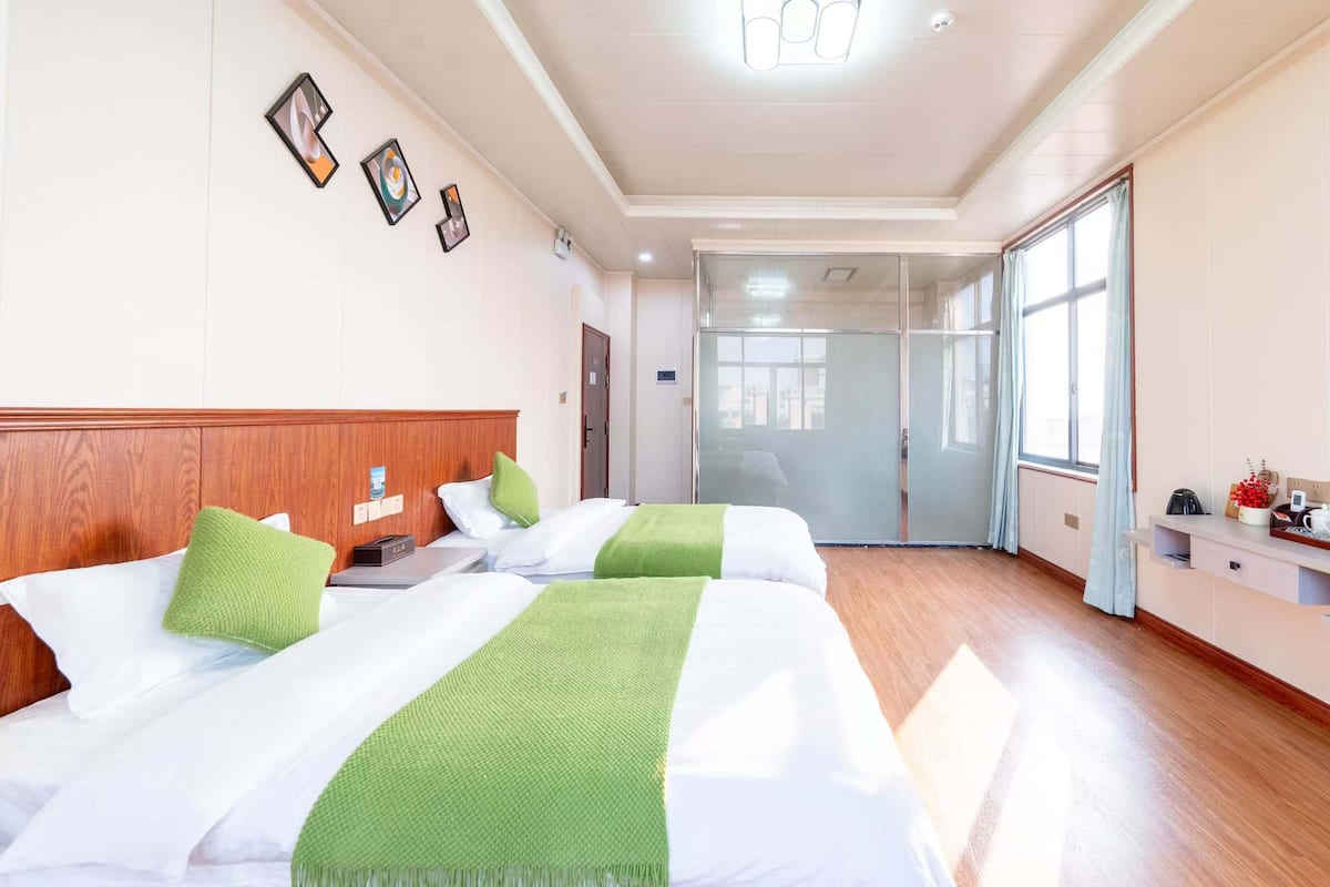 A hotel room with two beds, a wooden headboard, a nightstand, a desk, a chair, a window with curtains, and a door leading to a bathroom.