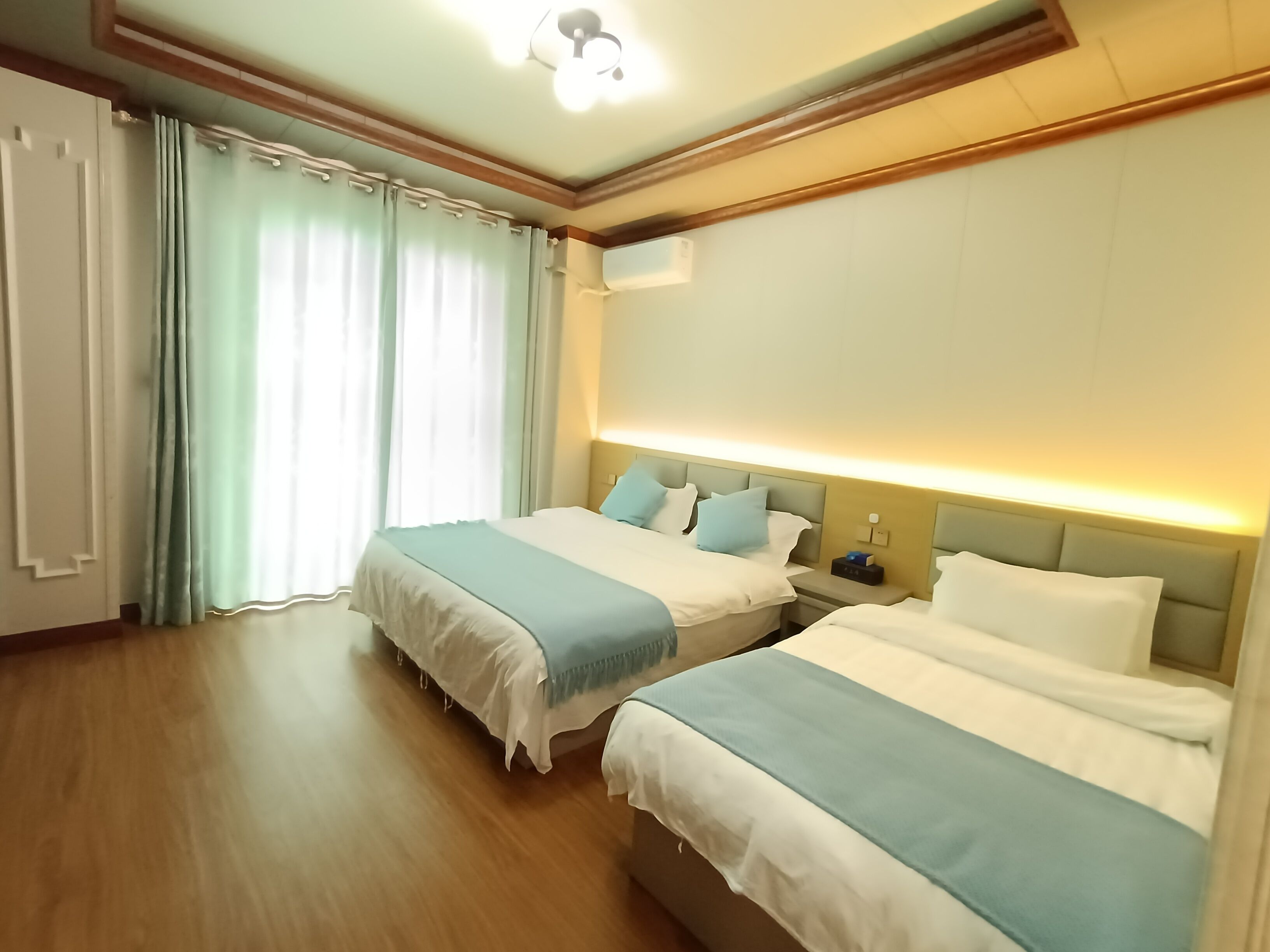 Habitación de hotel con dos camas, suelo de madera, ventilador de techo y una ventana grande con cortinas.
