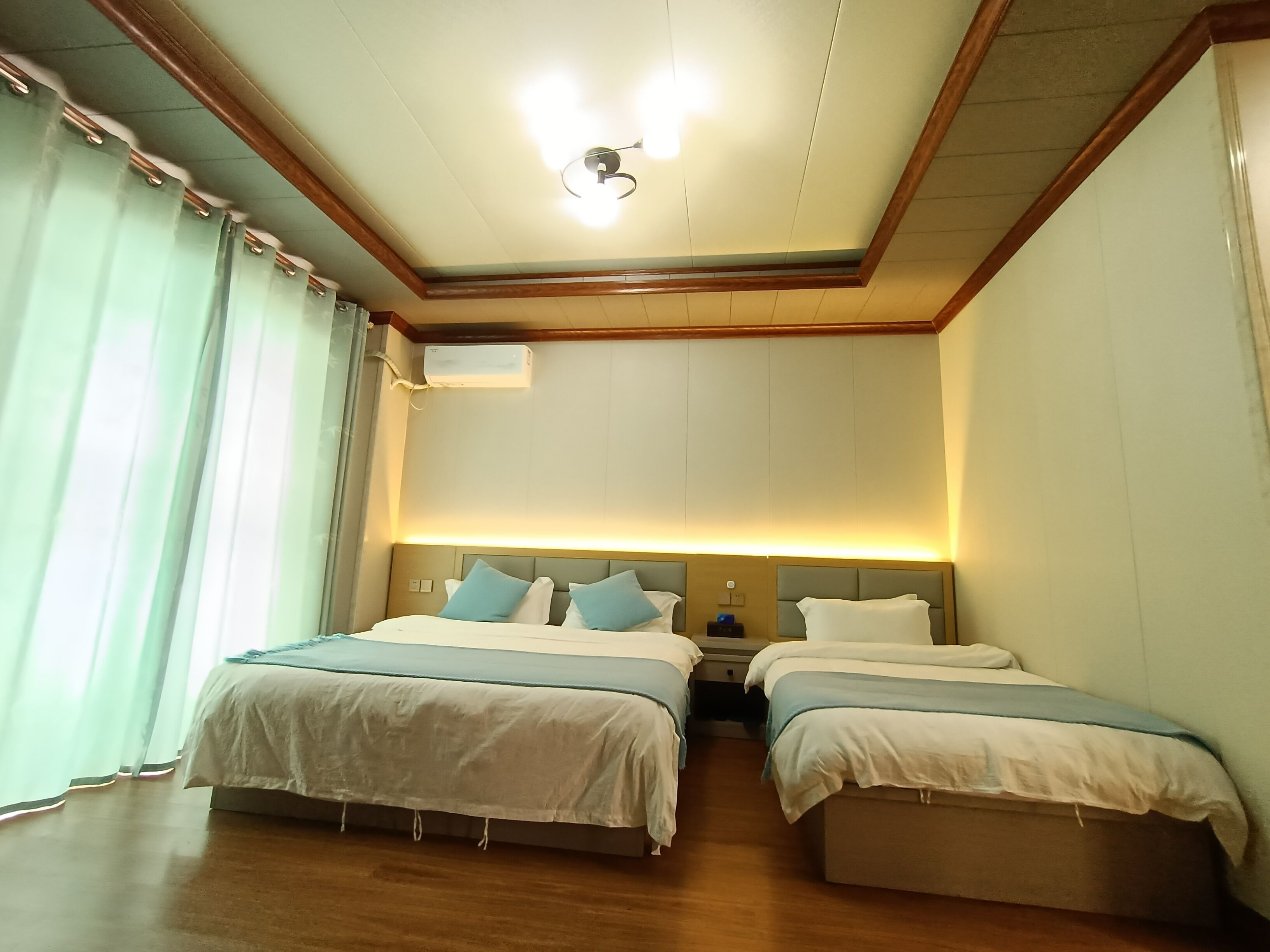 Habitación de hotel con dos camas, una lámpara de techo, un equipo de aire acondicionado y una mesita de noche con lámpara.