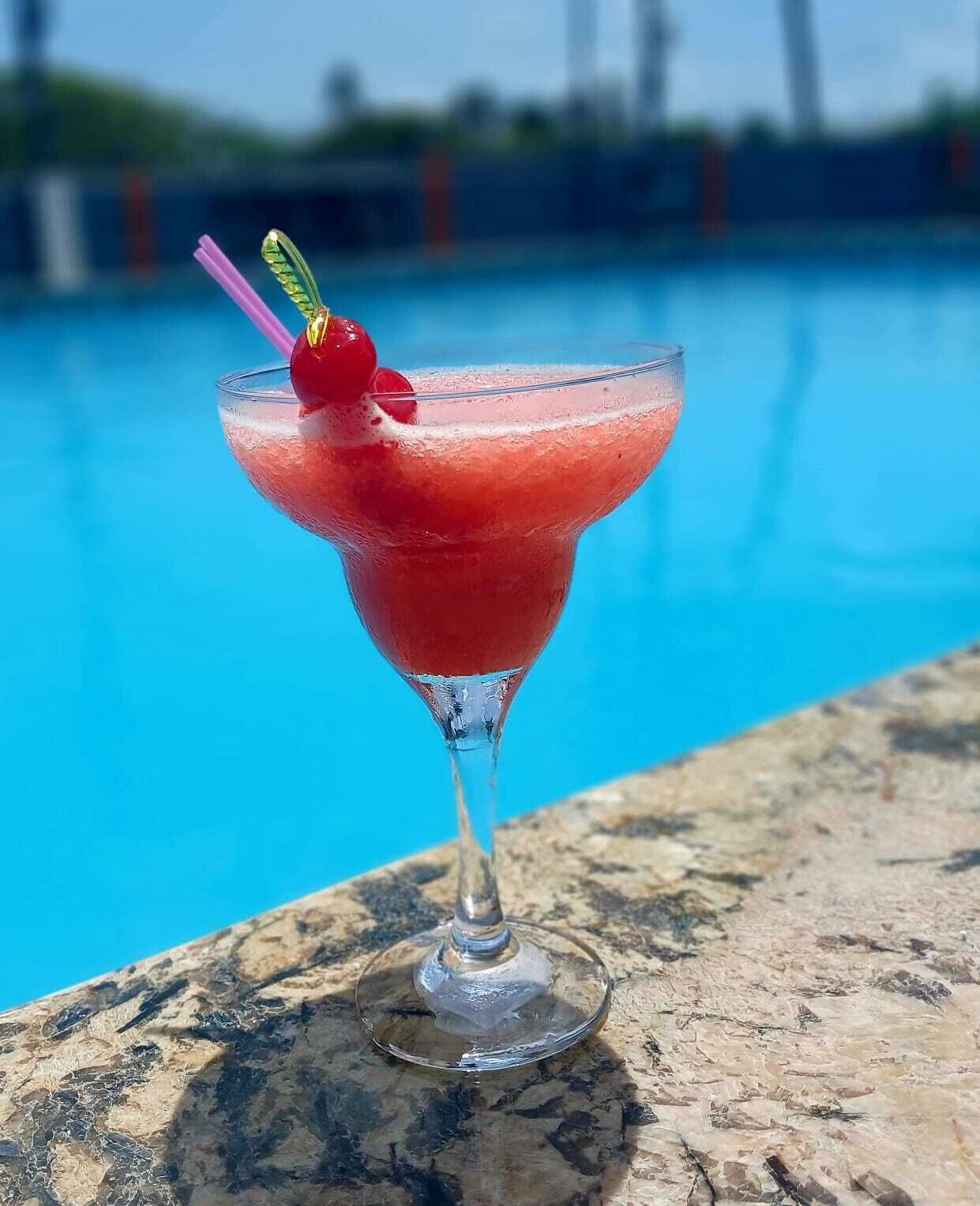 Ein Cocktail im Martini-Glas mit einer Kirsche am Spieß, auf einer Steinfläche neben einem Pool.