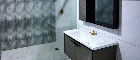 Un baño moderno con ducha envuelta en vidrio, un lavabo blanco con grifo dorado y un mueble de tocador de tonos oscuros.