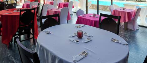 A restaurant with round tables covered in white and pink tablecloths, arranged in a spacious outdoor area with a pool view.
