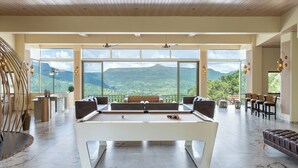A spacious room with a pool table, comfortable seating, and large windows offering a mountain view. - Villa 27 Amarja's Mountain Crest (Mawal)