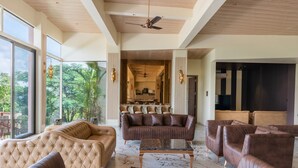 A spacious living area with leather sofas, a tufted armchair, and a marble coffee table. Large windows offer a view of lush greenery. - Villa 27 Amarja's Mountain Crest (Mawal)