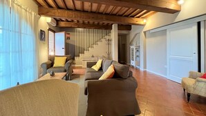 A spacious living room with a wooden ceiling, a staircase leading to another level, and a view of the outside through large windows. - Ludovica and Marina Villas with 2 pools (Lajatico)