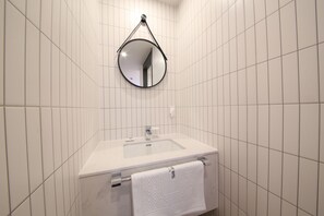 Un baño con paredes de azulejos blancos, un lavamanos blanco con un espejo encima y una toalla colgada en un perchero.