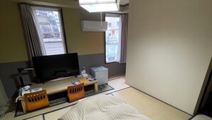 Una habitación de hotel con suelo de tatami, una cama, un televisor, un escritorio con computadora y una ventana con cortinas.