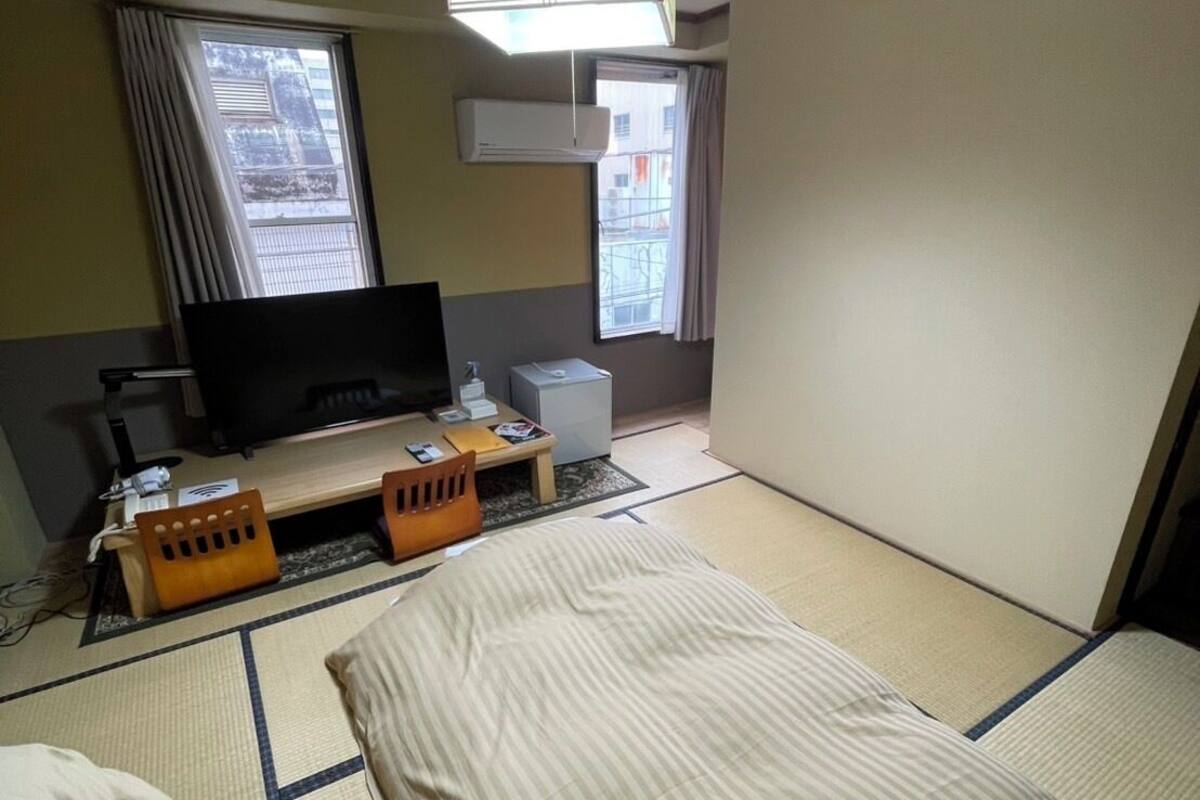 Una habitación de hotel con suelo de tatami, una cama, un televisor, un escritorio con computadora y una ventana con cortinas.