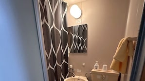 Un baño pequeño con un lavamanos blanco, un bidé y un inodoro. Hay un perchero con una toalla colgada, y una cortina estampada.