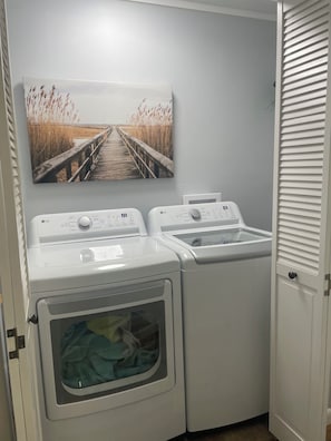 A laundry room with two front-loading LG washers and dryers, a wall-mounted picture of a beach, and a white door with a window. - Charming 2-bedroom house in quiet small town near Nashville (Ashland City)