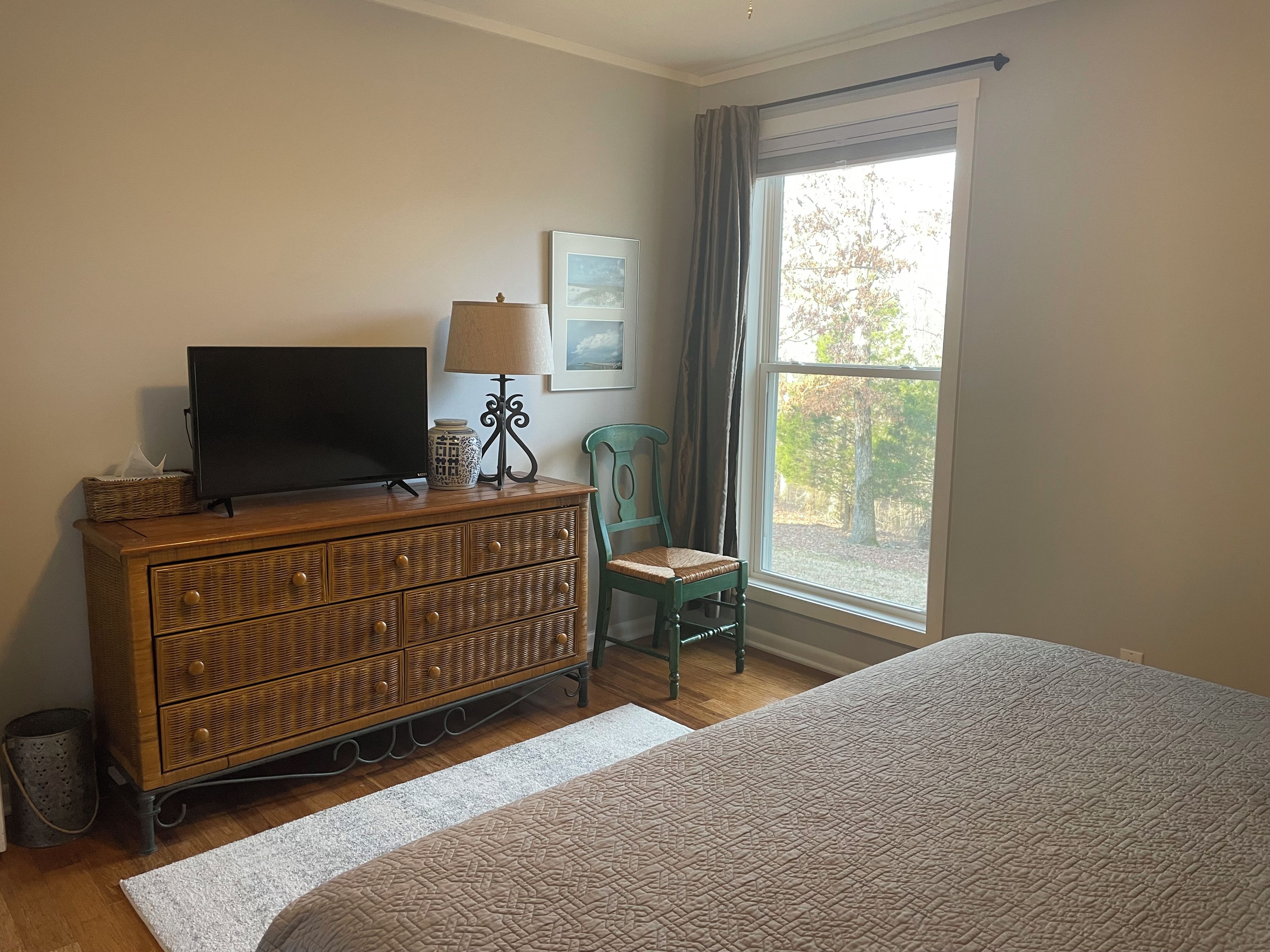 A bedroom with a bed, a wooden dresser, a television, a lamp, a chair, and a window with a view of trees.