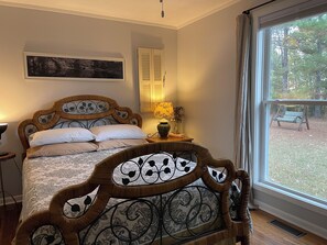 Ein Schlafzimmer mit Bett, Nachttisch, Lampe, Fenster mit Blick ins Freie und einem gerahmten Bild an der Wand.
