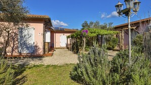 Un edificio de estilo mediterráneo tradicional con un camino de grava que lleva a una puerta, rodeado de exuberante vegetación y una pérgola con flores moradas.