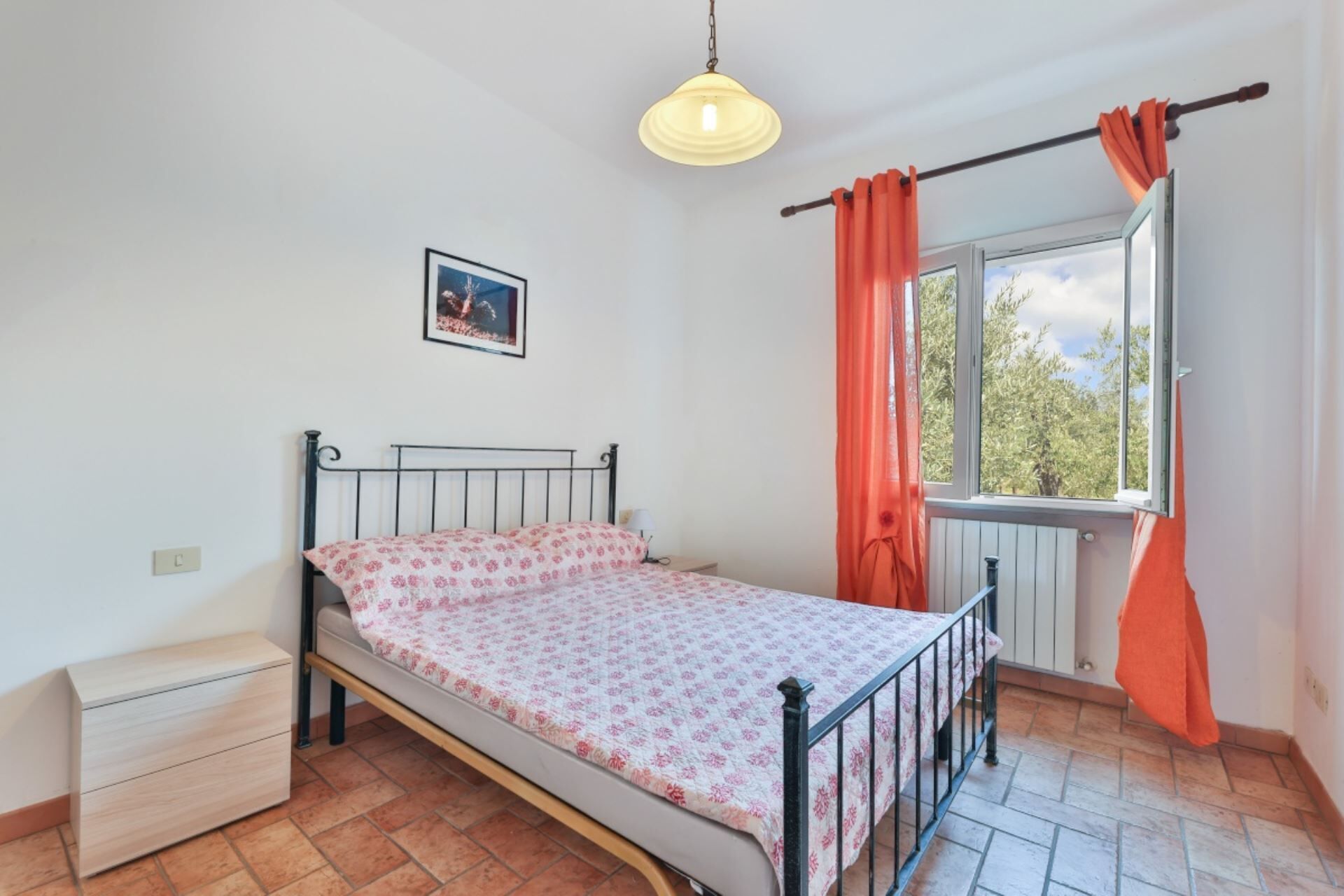 A bedroom with a bed, a nightstand, a dresser, a window with curtains, and a hanging light fixture.