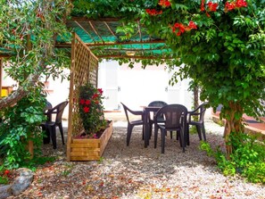 Un área de estar al aire libre con una pérgola, una jardinera de madera con flores y sillas de plástico.