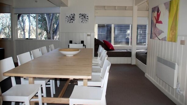 Un comedor moderno con una larga mesa de madera y sillas blancas, un banco con cojines, y vistas a árboles a través de amplias ventanas.