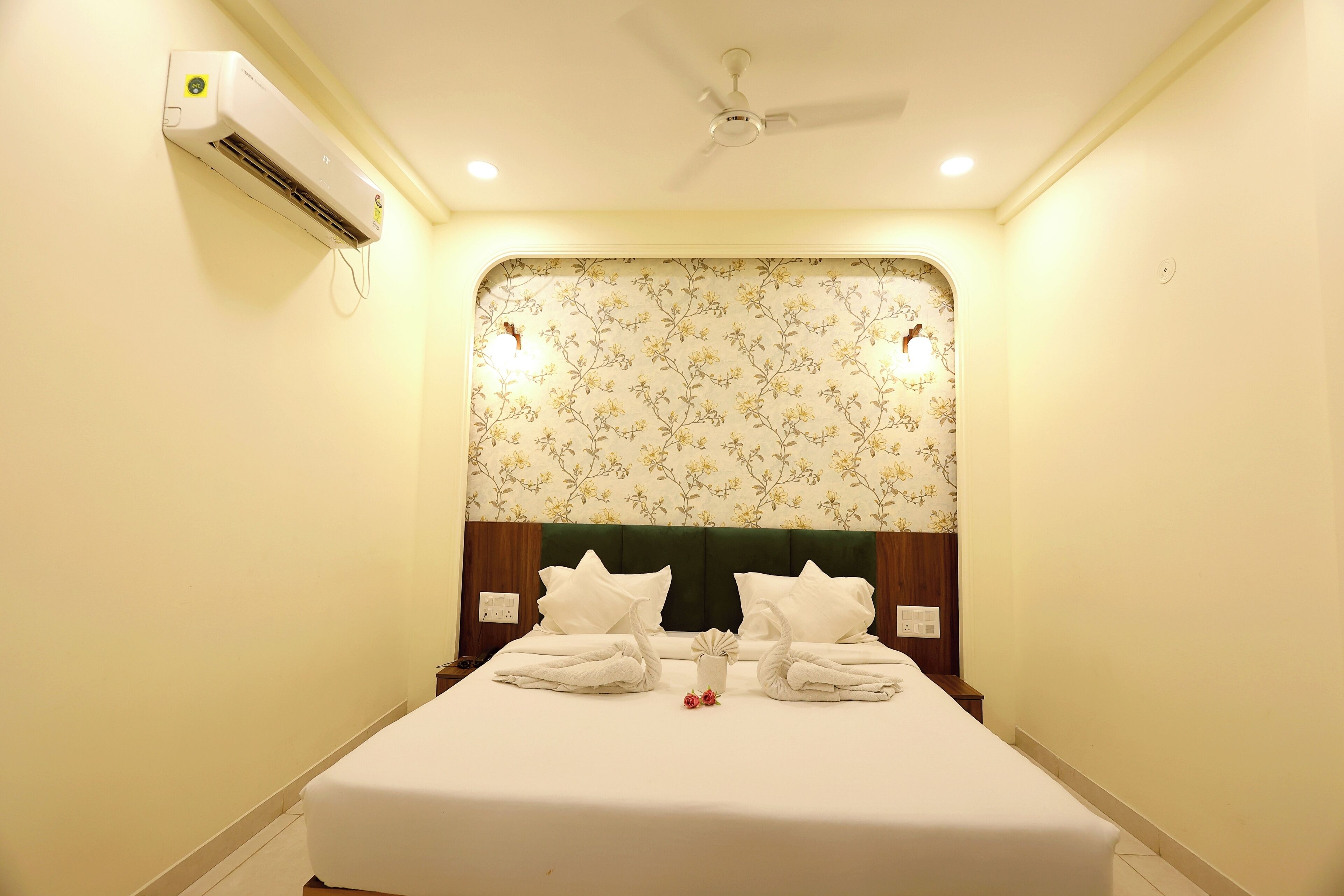 A hotel room with a bed, a ceiling fan, an air conditioning unit, floral wallpaper, and a nightstand with a lamp.
