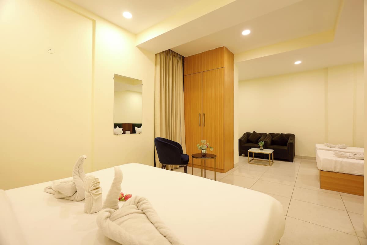 A hotel room with a large bed, a small table with a vase, a dark blue chair, a wooden wardrobe, a dark sofa, and a small white table.