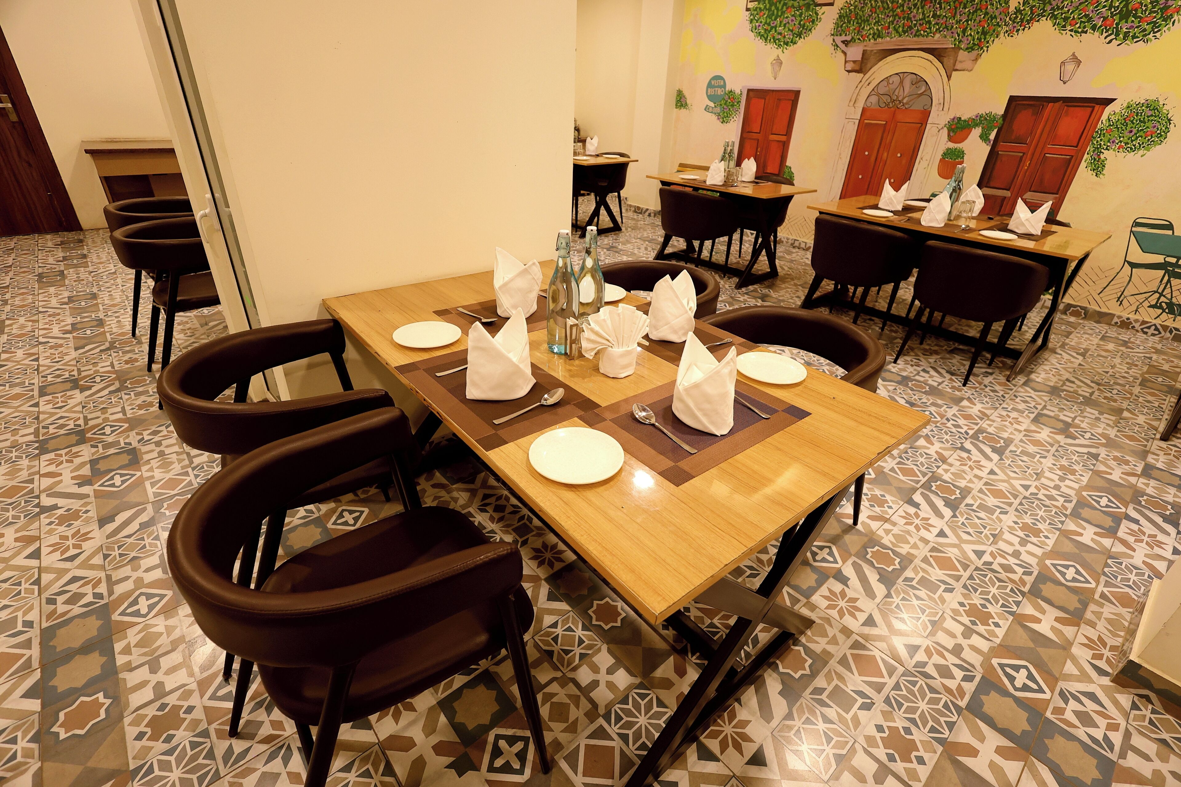 A dining area with a wooden table set for four, surrounded by chairs, with patterned floor tiles and a mural on the wall.