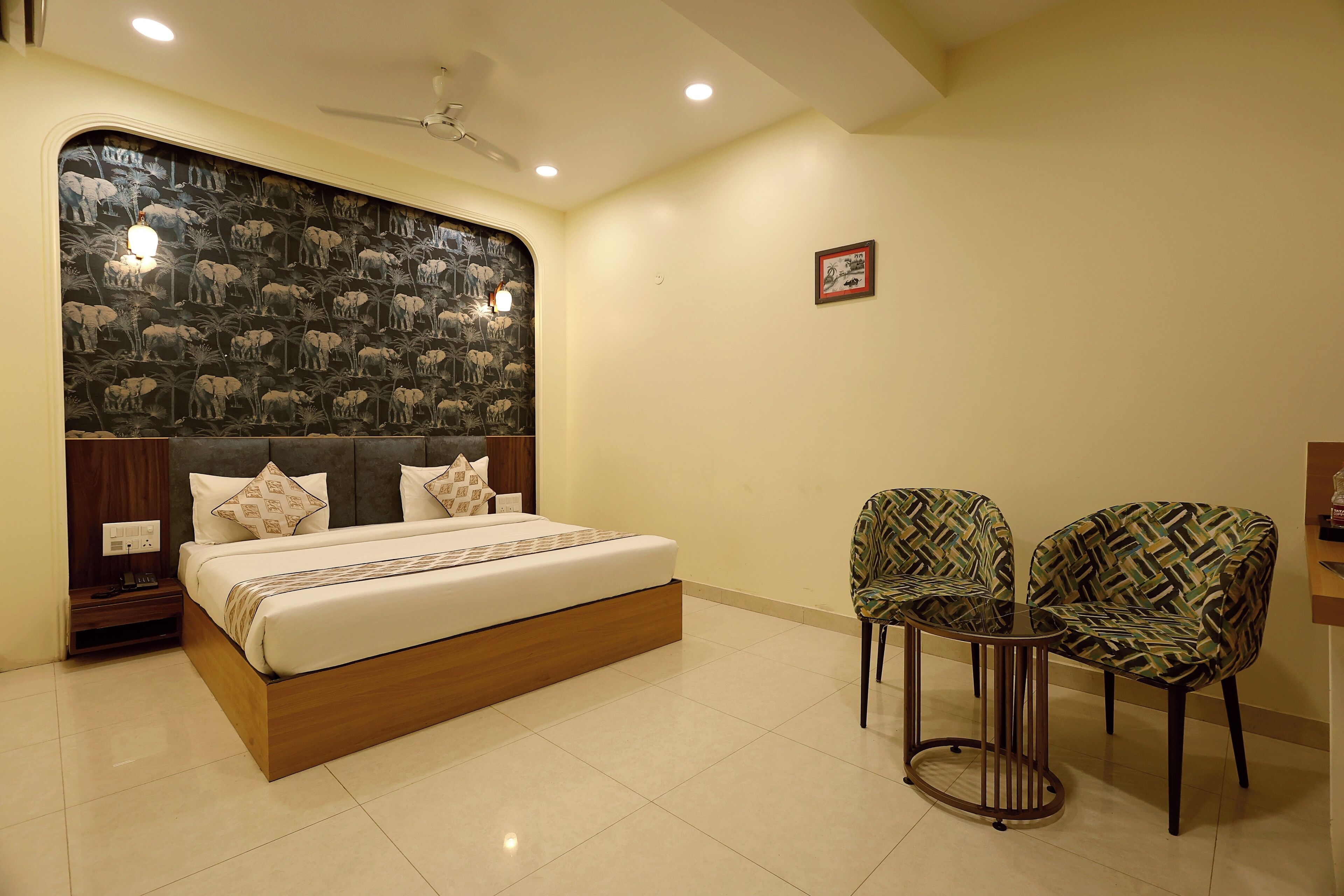 A bedroom with a bed, two chairs, a nightstand, and a wall with elephant wallpaper.