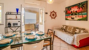 Un área de comedor con vista al mar, un timón de barco fijado a la pared y una pintura de atardecer tropical.