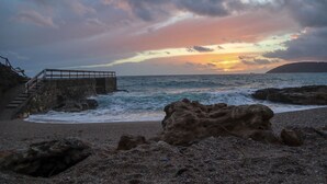 Una playa rocosa con una escalera que desciende hacia el agua, una gran formación rocosa en primer plano y un atardecer sobre el océano.