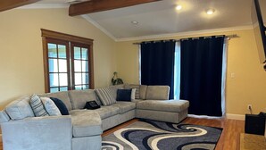 A living room with a sectional sofa, a dark blue rug, wooden flooring, a window with curtains, and a ceiling with exposed beams. - 3-bedroom Charming Home in the Hills of Pittsburg with hillside view & pool (Pittsburg)