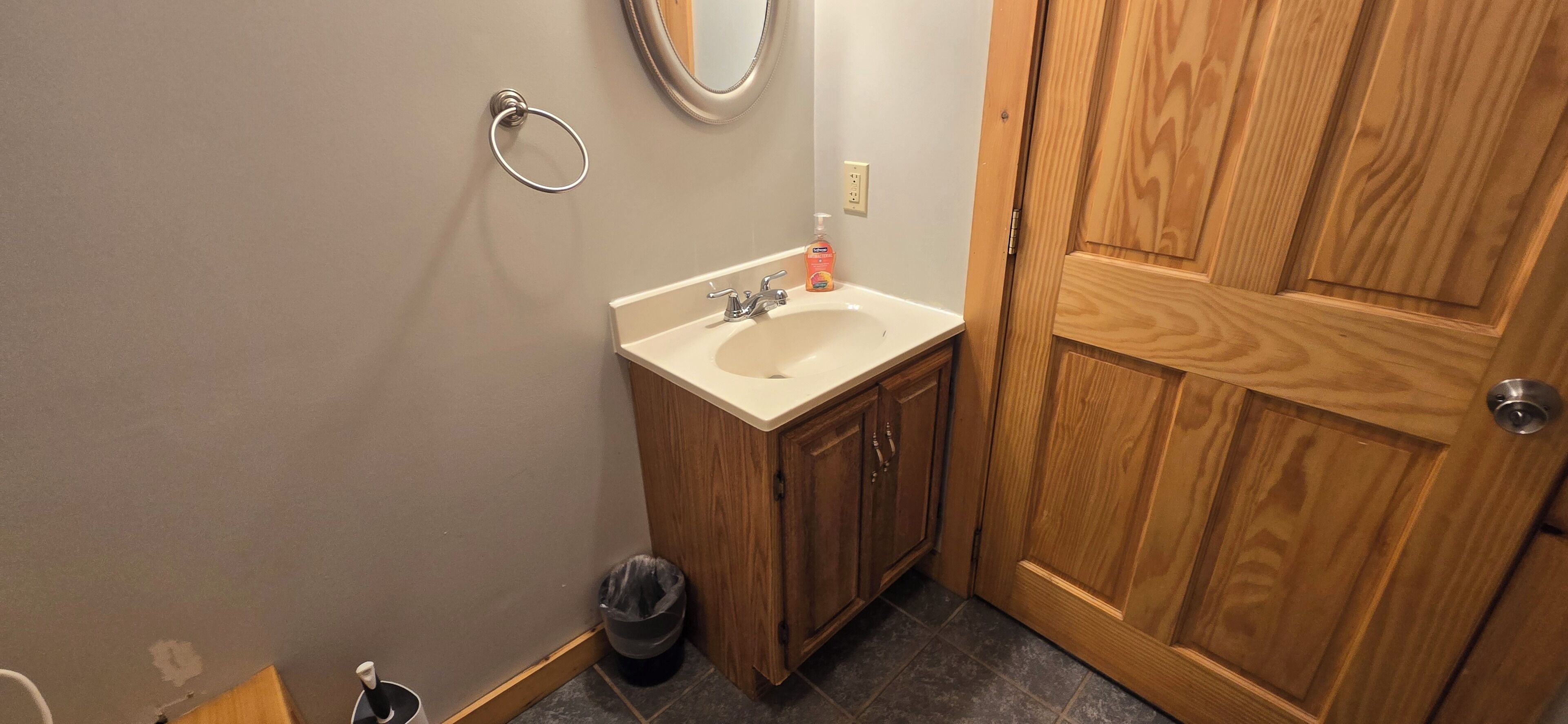 A small bathroom with a wooden door, a sink with a mirror above it, and a trash can.