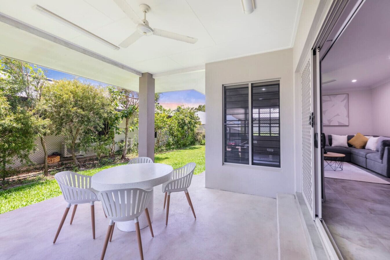 Une terrasse moderne avec une table ronde et des chaises, un ventilateur de plafond, et une vue sur un jardin et un bâtiment.