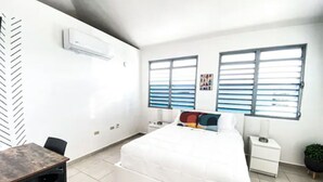 Ein modernes Schlafzimmer mit einem großen Bett, einem Nachttisch, einem Schreibtisch und einem Stuhl. Zwei Fenster mit Jalousien, eine Wand-Klimaanlage und ein kleiner Topfpflanze sind vorhanden.
