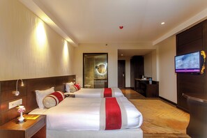 A hotel room with two beds, a TV, a desk, and a bathroom visible through a glass door.
