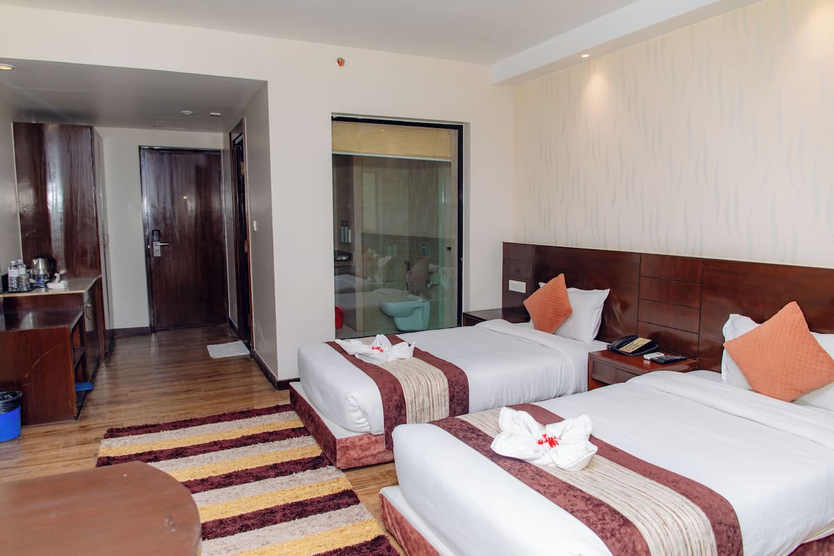 Habitación de hotel con dos camas, un escritorio y un baño visible a través de una puerta de cristal.