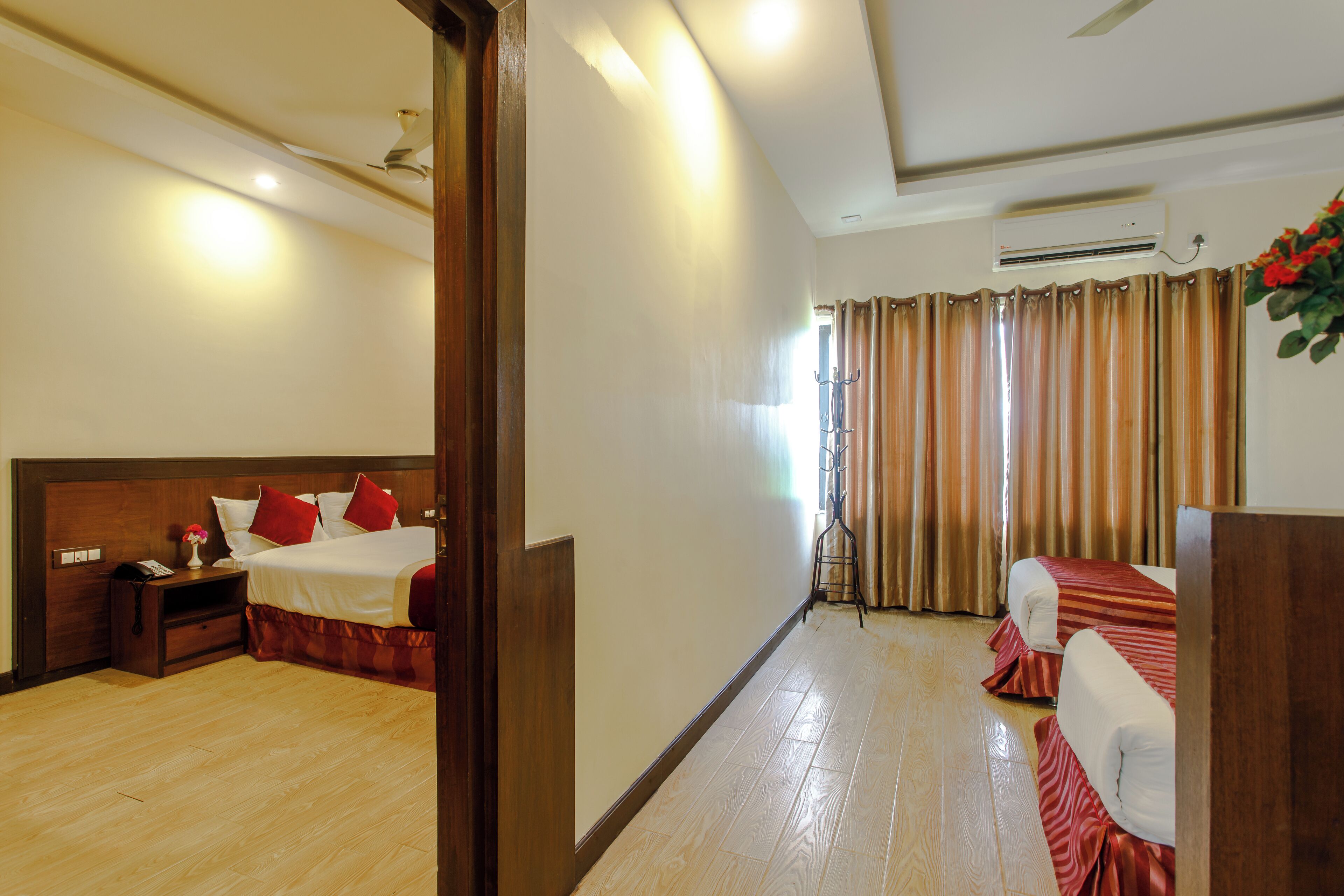 A hotel room with a wooden floor, a bed with red bedding, a bedside table with a lamp, and an air conditioning unit.